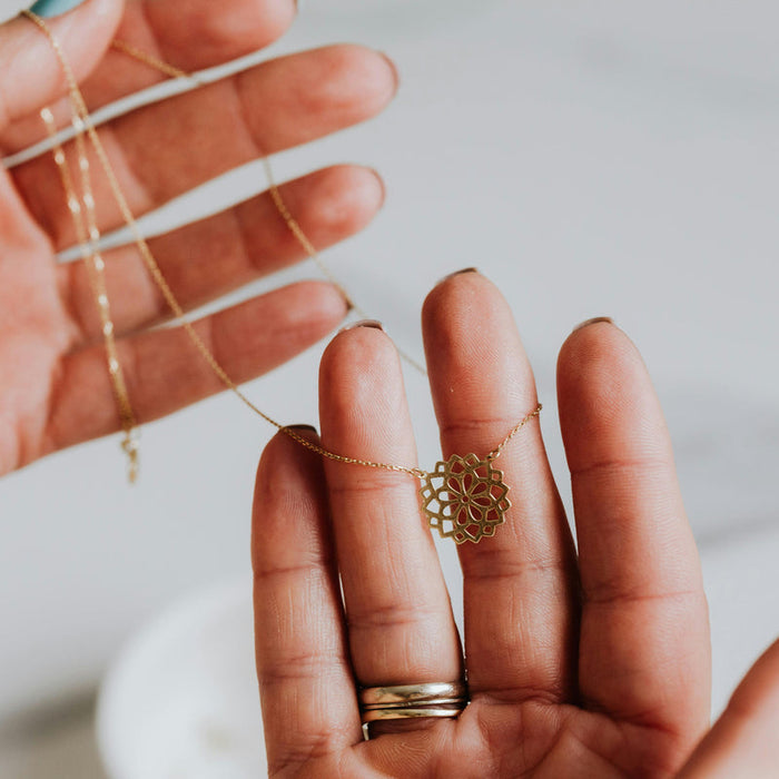 Hands holding a gold-colored floral necklace against a pale background; illustrative pendant styling photograph